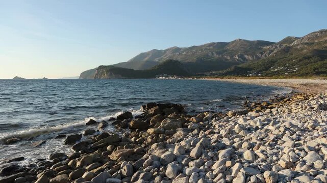 A pebble shoreline, gentle waves lapping against the stones, a tree-covered headland in the distance, and the afternoon sun.