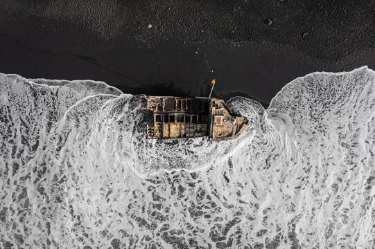 Aerial view of a boat stranded on the black sand beach of Pantai Wates Yeh Malet, surrounded by white sea foam waves in Kecamatan Karangasem, Bali, Indonesia.