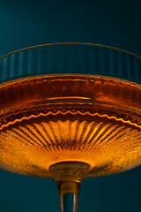 Naklejka na ściany i meble Extreme close-up macro of an amber cocktail in a textured, vintage crystal coupe glass with fine ribbed patterns and warm glowing light against a dark teal background.