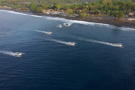 Aerial view of traditional jukung outrigger boats sailing on the blue sea near the black sand beach of Pantai Wates Yeh Malet in Kecamatan Karangasem, Bali, Indonesia.