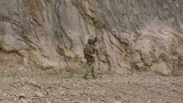 A special forces soldier in full combat gear advances cautiously through a dangerous, rocky valley, conducting a tactical patrol during a reconnaissance mission.