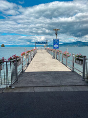 Fototapeta premium Lake geneva pier extending into calm waters under a dramatic cloudy sky, with blooming flowers lining the railings and distant mountains creating a serene travel destination scene