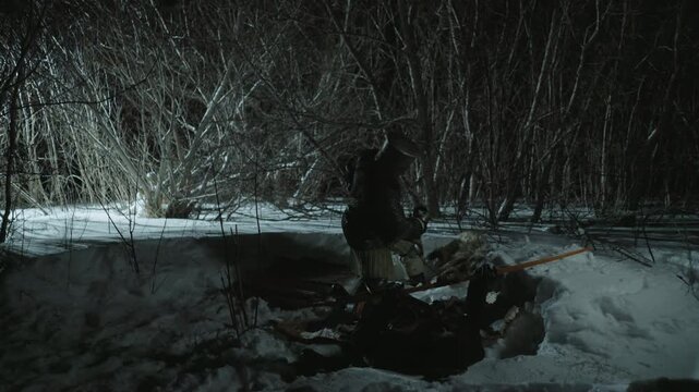 Armed figure in medieval armor stands over prone person at night within snowy dark forest. Character holds sword aloft and polearm suggesting hunt or combat. Tense scene with snow covered