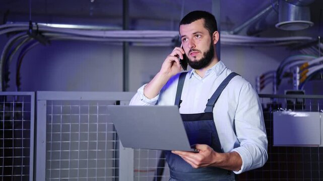 Dedicated technician with a beard, wearing work overalls and a light shirt, communicates on a smartphone while operating a laptop inside a busy server room, managing technical operations.