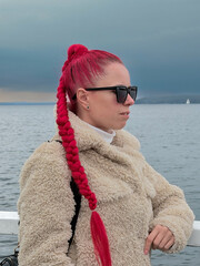 Close-up profile portrait of a woman with vibrant red hair braid wearing sunglasses near the water © Анастасия Константне