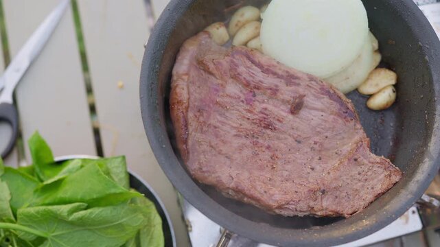 A large seasoned steak sizzles in a pan with fresh onion and garlic cloves, cooking on a portable stove for an outdoor barbecue or camping meal experience.
