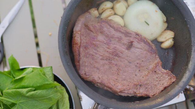 A large beef steak sears in a hot skillet with fresh garlic and onion, capturing the essence of outdoor culinary preparation for a gourmet camping meal.
