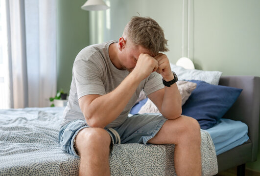 Stressed young man sitting on bed in morning holding his forehead due to severe migraine pain