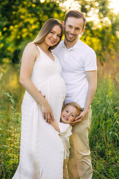 Happy pregnant family portrait: smiling couple with young son in summer meadow, maternity and parenthood moments