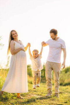 Pregnant mother and father playfully swing their young son outdoors at golden hour family portrait