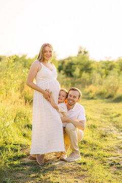 Pregnant mother with smiling husband and young son embracing in a sunlit summer meadow