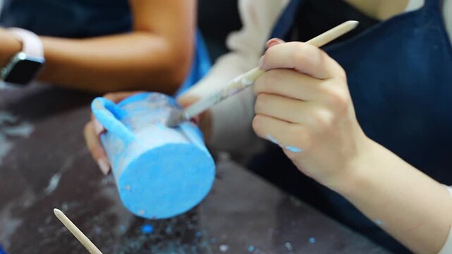 Woman applying blue glaze to handmade ceramic mug with brush. Pottery glazing process represents ceramic craft and handmade production
