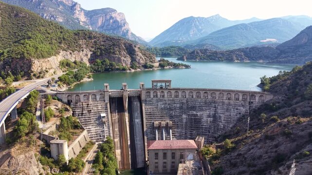 Aerial view of a massive concrete gravity dam with hydroelectric power station, turquoise reservoir, rocky mountain cliffs, and winding road in a scenic valley