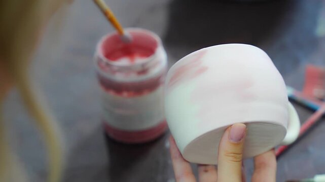 Woman applying glaze inside handmade ceramic mug with brush in pottery workshop. Pottery glazing process represents ceramic craft and handmade production