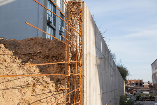 A close-up of steel rebar embedded in a concrete wall at a construction site. The retaining wall and the soil stand out against the backdrop of the building