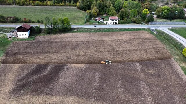 Aerial view of tractor tilling farm field. Rows of dark soil stretch across open land. Road runs beside the field, lined with trees. Houses dot the countryside in background. Farmer operates machine