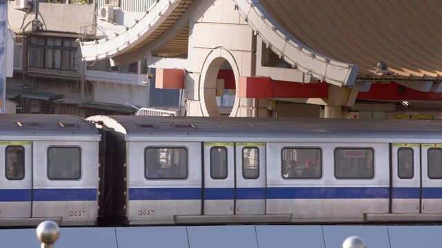 Slow motion follows a silver Taipei metro train passing below a traditional tiled roof, with blue stripes along the carriages and rounded metal posts in the foreground beneath the curved roof opening.