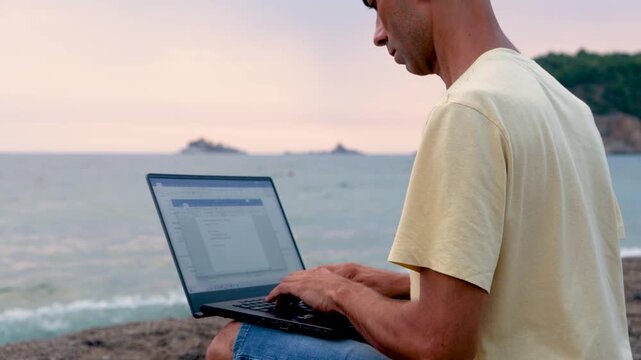 Freelancer using laptop on rocky beach during peaceful sunset by sea