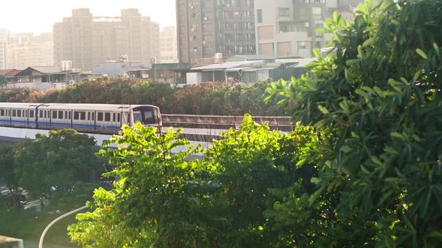 A metro train enters from the left and moves right along an elevated Taipei track in slow motion, beyond sunlit leaves and branches with closely spaced apartment buildings forming the city background.
