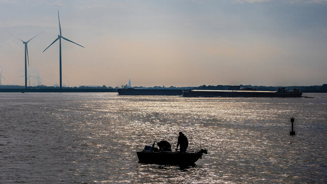 Fisherman on a boat with wind turbines, barges, and sunset