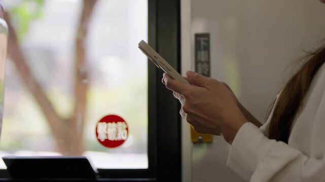 A masked passenger in a white jacket holds a smartphone upright beside a bus window in Taipei, seen in slow motion with hands near the screen and the face cropped above the mask inside the city bus.
