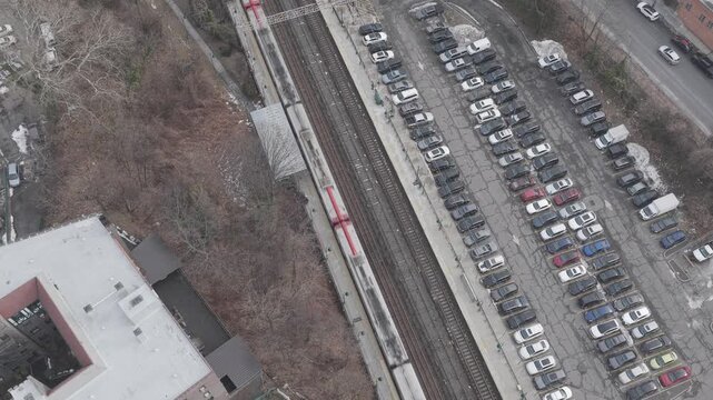 Top down drone glide above parked cars beside railway tracks. Rows of parked vehicles run parallel to tracks and a station platform in a steep overhead composition.