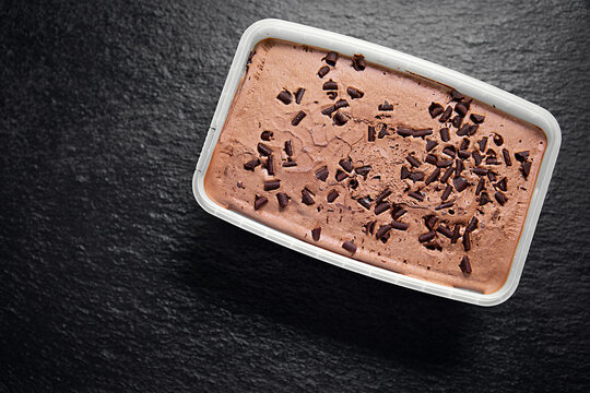 Chocolate ice cream with chocolate chips in a plastic container, photographed from above on a dark textured background.