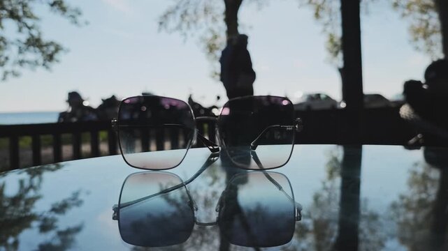 Pair of sunglasses sitting on a reflective table outdoors. In the blurred background, people are walking in a park near the sea