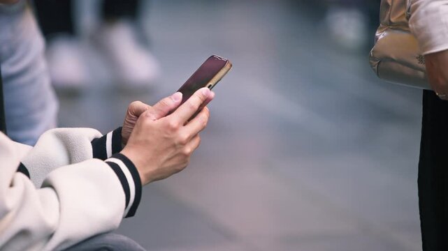A seated person in a white jacket uses a smartphone in slow motion beside a Taipei sidewalk, with striped cuffs around the hands and passing pedestrians appearing as shapes beyond the cropped torso.