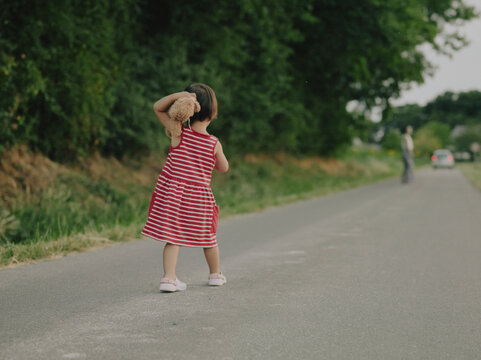 Young girl in a red striped dress holding a teddy bear, walking away along a country road in rural Pays de la Loire, France.