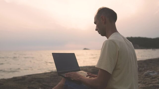 Man typing on laptop at coastal beach during tranquil sunset hours