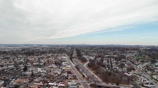Wide aerial pan across divided highway and winter neighborhoods. Multiple lanes curve through a dense pattern of rooftops beneath a pale cloud streaked sky. Filmed in Queens NYC.