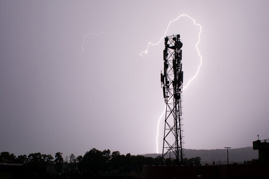 A dramatic lightning bolt strikes a cell tower silhouetted against a stormy sky, capturing the intensity of a thunderstorm and the power of nature.