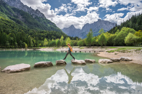 Woman hiker walking on stones across Jasna Lake in Slovenia. Alpine autumn landscape with sporty girl, turquoise water reflection, sky, clouds, green trees forest, yellow backpack and mountain view