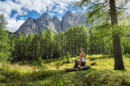 Woman hiker sitting on wooden bench in autumn forest in Slovenia. Alpine landscape with larch trees, sky, clouds, rocky mountain peaks, hiking trail, green meadow and bright sunny day view