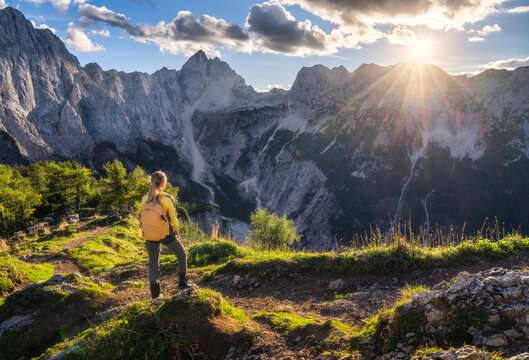Woman hiker with yellow backpack standing on trail in Slovenia. Alpine autumn landscape with rocky mountain peaks, sky, clouds, green trees forest, evening sunset light. Trekking in Alps. Autumn