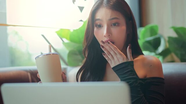 Surprised young Asian woman reacting with positive excitement while looking at laptop screen, covering mouth with hand and holding takeaway drink, unexpected good news, pleasant surprise