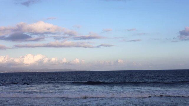 Ocean waves roll toward calm shoreline beneath wide blue sky. Gentle surf breaks in parallel lines, offering coastal view relevant to shoreline erosion and changing marine environments.