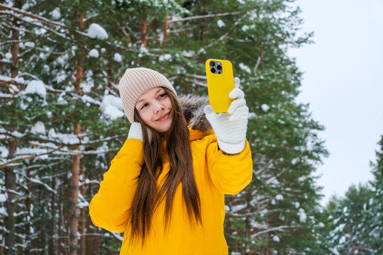 Young woman taking selfie with smartphone in snowy pine forest while wearing bright yellow winter jacket and knitted hat. Winter travel, social media and outdoor leisure concept.