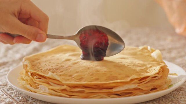 Human hand uses spoon to spread sweet berry jam on top of hot homemade pancakes stack during slow motion. Cinematic morning breakfast video footage provides tasty asset for design element.
