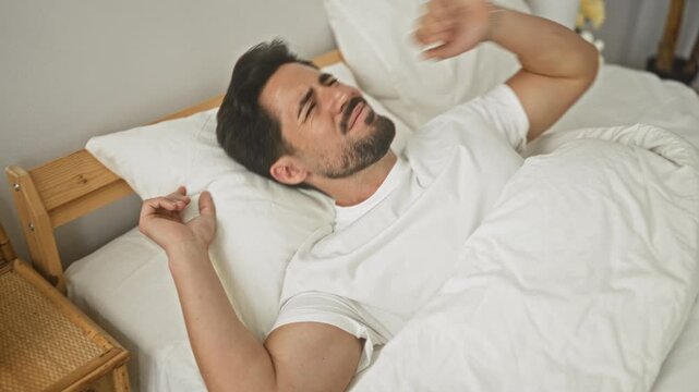 Man on bed grimacing with hand to head and pulling duvet over face in a building; morning discomfort.