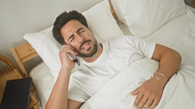 Man in bed holding smartphone to ear, hand on chest, wincing and squinting inside a residential building near bedside table; late night concern.