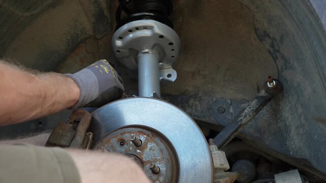 Mechanic hands in gloves installing a new silver front suspension strut shock absorber with coil spring into a car wheel well during a repair process in a workshop, close up of car maintenance. 
