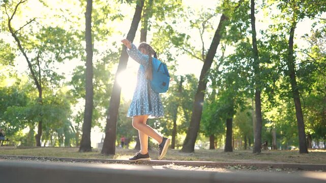 little girl walking along the curb. concept of happy childhood and loving family. a schoolgirl in a dress and with a blue backpack walks along the curb, keeps lifestyle her balance