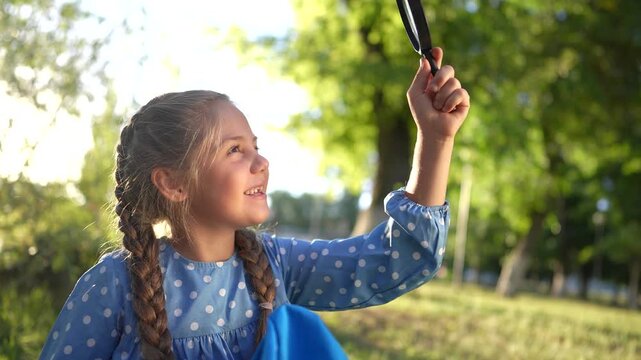 the girl holds magnifying glass in her hands. gaining knowledge and curiosity concept. a lifestyle schoolgirl girl with a black magnifying glass is examining something, park on the background