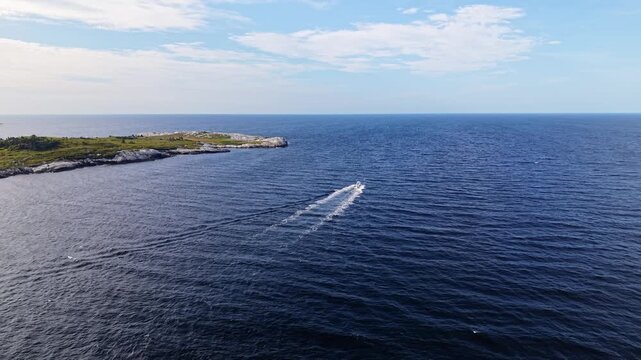 Aerial view of a white fishing boat with an outboard motor speeding through Atlantic Ocean waves at sunrise, passing the rugged rocky coastline of Prospect, Nova Scotia, Canada.
