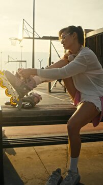 Sportive young woman pulls shoe strings on heavy inline skates. Golden sunset rays illuminate urban training ground while fit rider gets ready for fast rolling across city park.