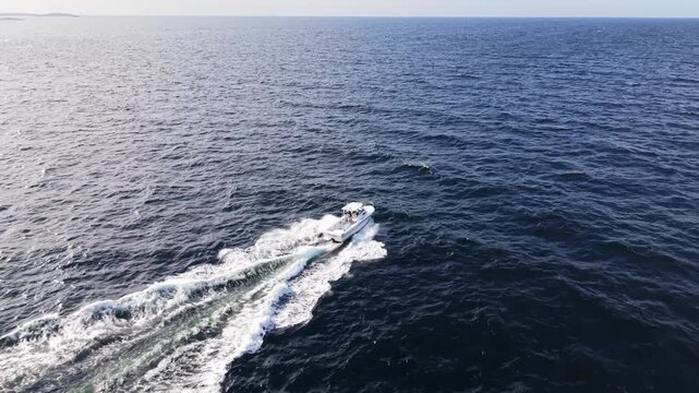 Aerial view of a white fishing boat speeding across the Atlantic Ocean waves near the rocky coastline of Prospect, Nova Scotia, Canada, leaving a long wake behind.