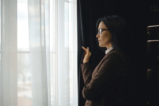 A businesswoman stands by a window in a modern office, gazing thoughtfully outside. She wears glasses and a stylish outfit, with soft natural light illuminating the room