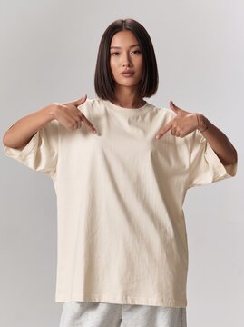 Woman wearing an oversized white t-shirt while pointing towards her chest against a plain studio background.